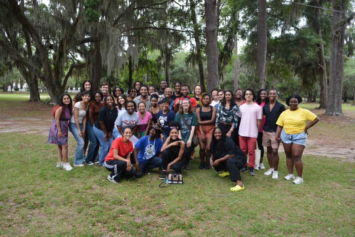 A group of people pose together outdoors under large trees, standing and kneeling on grass with a wooded background, gathered for a Penn Center partnership event with the Willson Center.