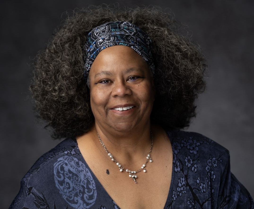 Smiling woman with gray curly hair, Artist-in-Residence at Penn Center, wears a patterned blue headband and matching top, posing against a dark gray background.